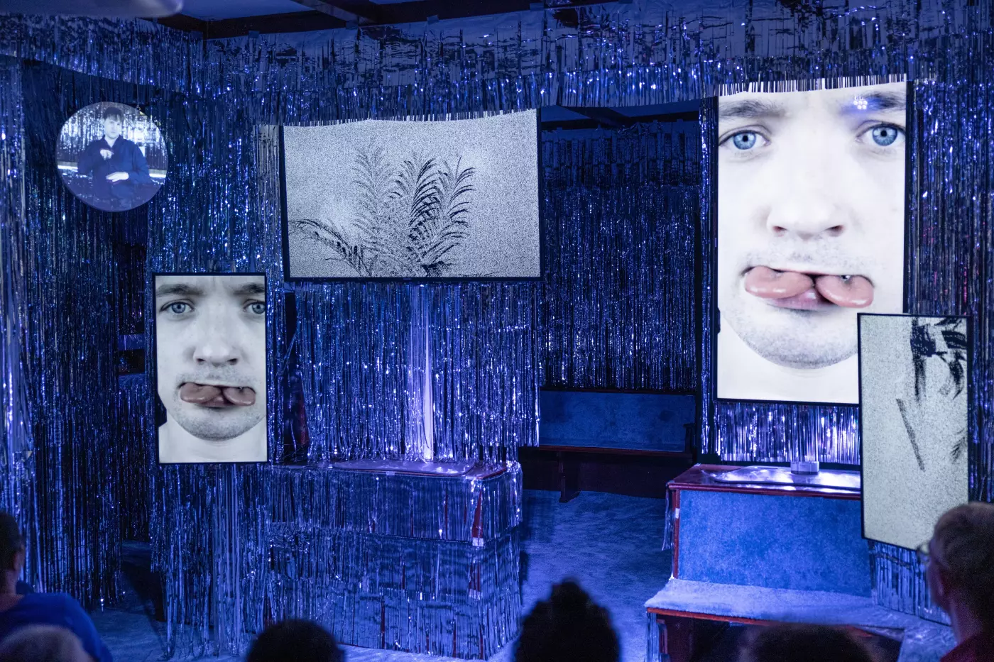 Eine leere Bühne und vier Bildschirme, auf denen das Gesicht des Schauspielers mit zwei Zungen gezeigt wird.